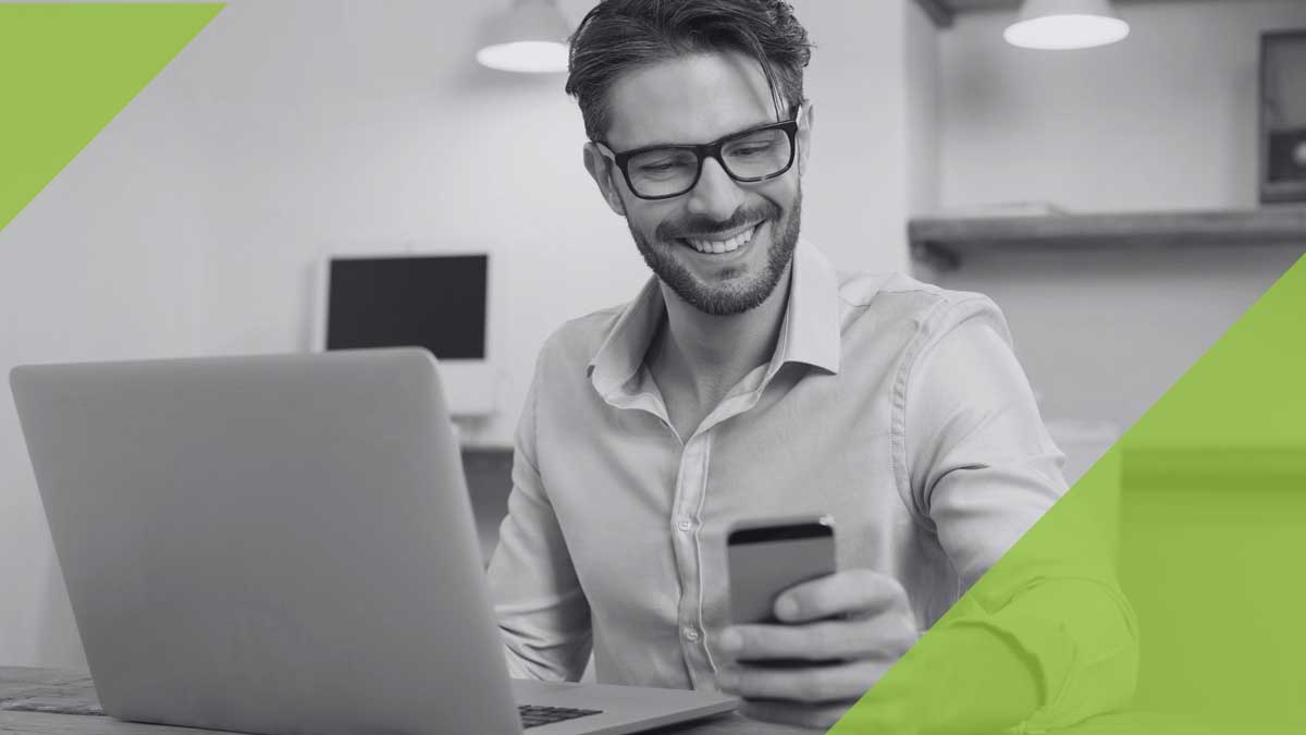 Mann vorm Laptop, der lächelnd auf sein Smartphone schaut